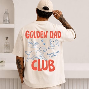 May include: Cream-colored t-shirt with the words "GOLDEN DAD CLUB" in red and blue. The shirt features illustrations of a dog, a dog bowl, and paw prints. The text "happier with a golden" is also included.