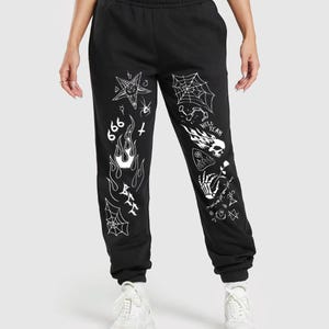May include: Black sweatpants with white graphic designs including a pentagram, spiderwebs, flames, and the number 666. The sweatpants have a drawstring waist and elastic cuffs. The model is wearing white sneakers.