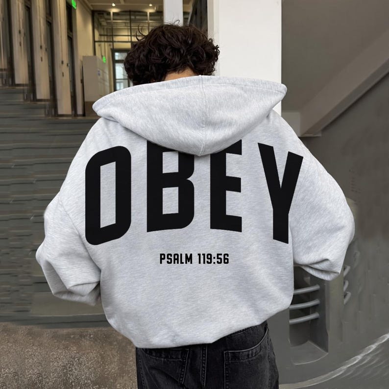 May include: A pale grey hoodie featuring the word "OBEY" in large black capital letters on the back. Underneath, the text "PSALM 119:56" is printed in black. The hoodie has a hood and long sleeves.