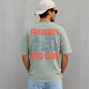 May include: Sage green t-shirt with the words "FRENCHIES" and "DAD CLUB" in orange block letters. The shirt features blue illustrations of French bulldogs and the phrase "happier with a french bulldog."