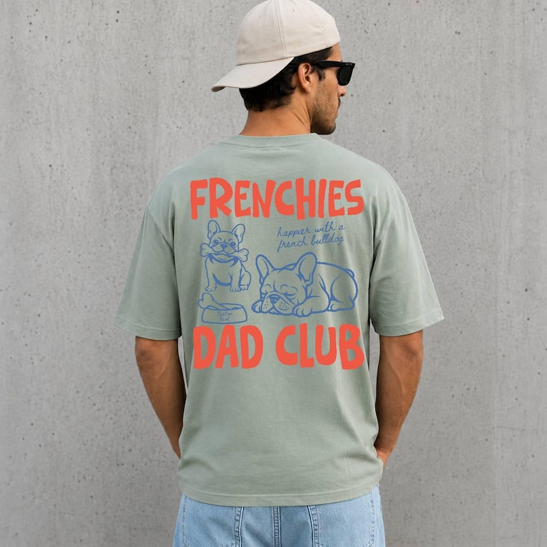 May include: Sage green t-shirt with the words "FRENCHIES" and "DAD CLUB" in orange block letters. The shirt features blue illustrations of French bulldogs and the phrase "happier with a french bulldog."