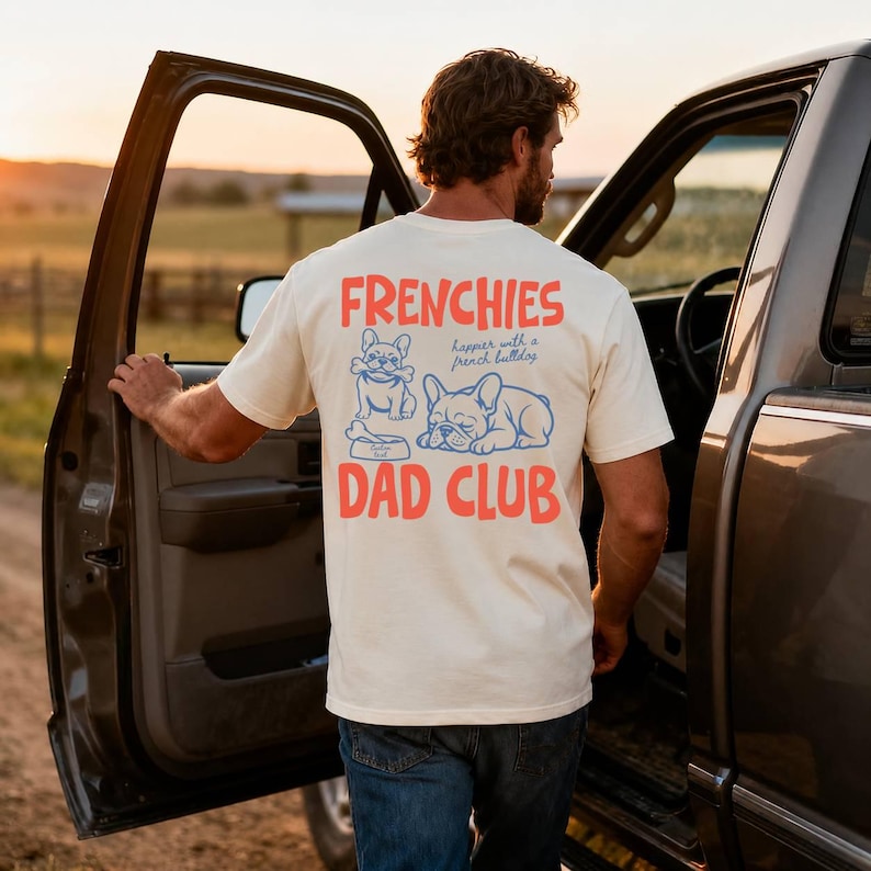 May include: Cream-colored t-shirt with the text "FRENCHIES DAD CLUB" in red and blue. The shirt features illustrations of two French Bulldogs. The person is standing next to a truck with the door open.