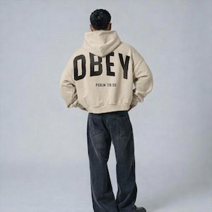 May include: A beige hoodie featuring the word "OBEY" in large black letters on the back, with "PSALM 119:56" underneath. The hoodie is worn with dark wash denim jeans. The person is seen from the back.