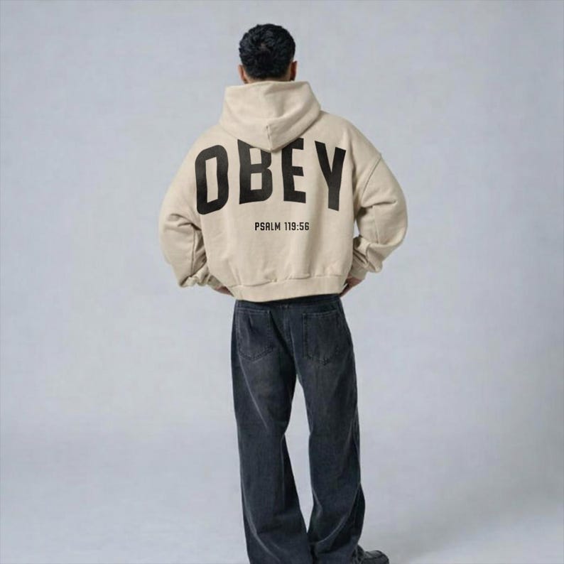 May include: A beige hoodie featuring the word "OBEY" in large black letters on the back, with "PSALM 119:56" underneath. The hoodie is worn with dark wash denim jeans. The person is seen from the back.