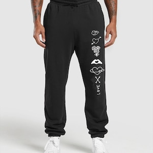 May include: Black sweatpants with a drawstring waist and white graphic design. The design includes hearts, arrows, roses, a mouth, a planet, and the word "LOVE" with an "X" symbol. The sweatpants are paired with white sneakers.