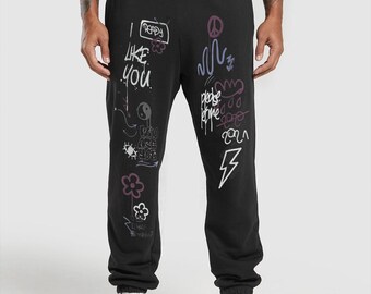 May include: Black sweatpants with a drawstring waist and white sneakers. The sweatpants feature graffiti-style drawings and text, including the phrases "I like you" and "please forgive". Other graphics include flowers, a peace sign, and a lightning bolt.