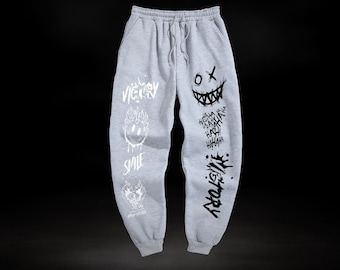 May include: Light grey sweatpants with a drawstring waist and pockets. The pants feature black graphic designs including a smiling face, the word "Victory", and the word "Smile".