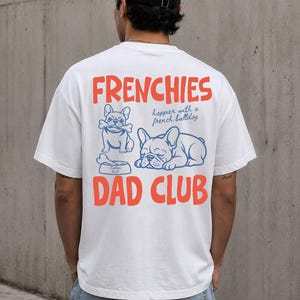 May include: White t-shirt with the words "FRENCHIES" and "DAD CLUB" in orange, and two blue French bulldog illustrations. The shirt also has the text "happier with a french bulldog".
