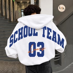 Pode incluir: Um moletom com capuz branco apresentando o texto SCHOOL TEAM em letras azuis grandes nas costas. Abaixo do texto, o número 03 está impresso em azul, com o nome Lewis escrito em letra cursiva laranja sobreposta ao número.