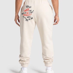 May include: Cream-colored sweatpants with elastic cuffs. The back of the pants features the phrases "HA! HA! WHY SO SERIOUS? HA HA HA HA!" in a mix of red and black graffiti-style lettering. The pants are made of a soft material.