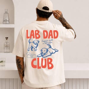 May include: Cream-coloured t-shirt featuring the text "LAB DAD CLUB" in red, accompanied by two blue Labrador illustrations. The shirt also includes the phrase "happier with a labrador".