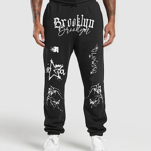 May include: Black sweatpants with white graphics and text. The word "Brooklyn" is written across the top in a gothic font. Other graphics include a star, a flame, and the word "Glowing". The sweatpants have a drawstring waist.