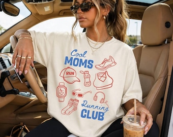 May include: Cream-colored t-shirt with the text "Cool Moms Running Club" in blue and red. The shirt features running-themed illustrations, including a shoe, hat, and water bottle. The person is wearing black leggings and sunglasses.