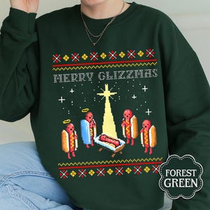 May include: Dark green sweatshirt with the words "MERRY GLIZZMAS" and a pixelated Christmas scene featuring hot dogs. The design includes a star, angels, and a baby hot dog in a manger. The sweatshirt has a festive, holiday theme.