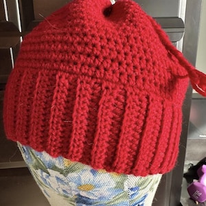 Pode incluir: Um chapéu vermelho feito à mão, de crochê, com uma aba canelada e um topo pontudo. O chapéu é exibido em uma forma com estampa floral. A textura do chapéu é visível, mostrando os pontos apertados e a espessura do fio. Uma borla pende do topo.