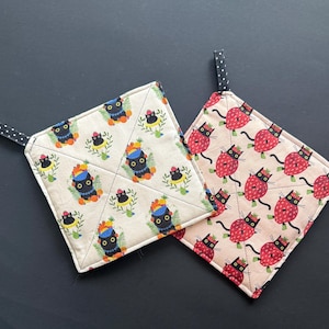 May include: Two square quilted pot holders with black fabric hanging loops. One features a cream background with a repeating pattern of black cats wearing colorful fruit hats. The other has a light pink background with black cats shaped like strawberries.