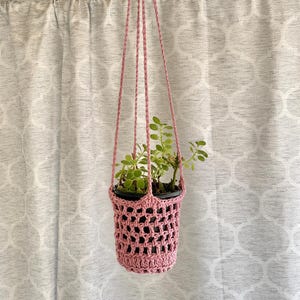 May include: A pink crocheted plant hanger with a potted plant inside. The plant hanger is suspended by four pink cords. The plant has green leaves. The background is a white curtain with a repeating pattern.