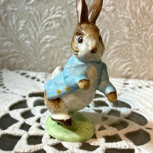 Puede incluir: Una figura de cerámica de un conejo de pie sobre una base verde. El conejo tiene pelaje marrón, detalles blancos y lleva una chaqueta azul claro con botones amarillos. La figura se muestra en una postura erguida y alerta con las orejas hacia arriba.