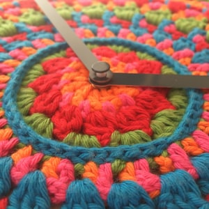 May include: A colorful crocheted clock face with black clock hands. The clock face is made of a variety of colors, including red, orange, yellow, green, blue, and purple.