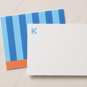 Può includere: Un biglietto bianco con la lettera K stampata in inchiostro blu, sovrapposto a una busta a righe azzurre e blu scuro con una fascia arancione sul bordo inferiore. Il set di cancelleria è un prodotto cartaceo piatto e rettangolare.
