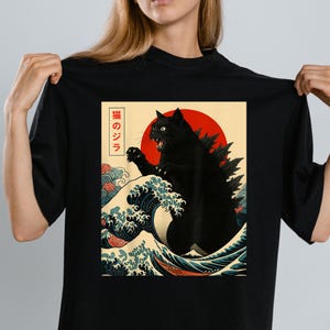 May include: Black t-shirt featuring a graphic of a black cat in a Godzilla-like pose, set against a red sun and ocean waves. The design includes Japanese text. The shirt is a crew neck.