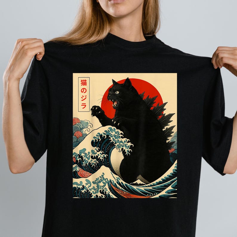 May include: Black t-shirt featuring a graphic of a black cat in a Godzilla-like pose, set against a red sun and ocean waves. The design includes Japanese text. The shirt is a crew neck.