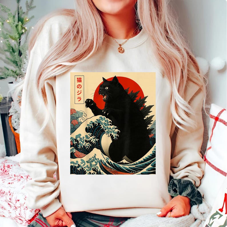 May include: Cream-colored sweatshirt featuring a graphic of a black cat with a Godzilla-like dorsal fin, set against a red sun and a stylized ocean wave design. Japanese text is present. The design is reminiscent of traditional Japanese art.