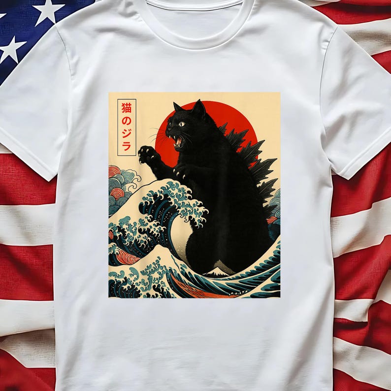 May include: White t-shirt featuring a black cat in a Godzilla-like pose, set against a red sun and ocean waves. The design includes Japanese text. The cat has a spiky dorsal fin. The waves are blue, white, and red.