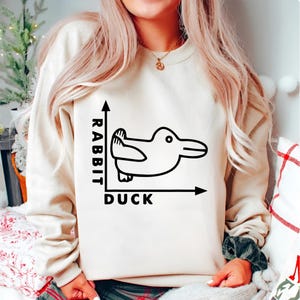 Może przedstawiać: Beżowa bluza z czarną grafiką przedstawiającą wykres iluzji kaczki-królika. Wykres ma słowa "Rabbit" i "Duck" oznaczające osie. Obraz kaczki-królika znajduje się na środku wykresu.