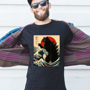 May include: Black t-shirt featuring a stylized illustration of a black cat in the style of a Japanese woodblock print. The design includes a red sun, ocean waves, and Japanese text. The cat has a Godzilla-like appearance.