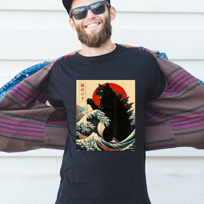 May include: Black t-shirt featuring a stylized illustration of a black cat in the style of a Japanese woodblock print. The design includes a red sun, ocean waves, and Japanese text. The cat has a Godzilla-like appearance.