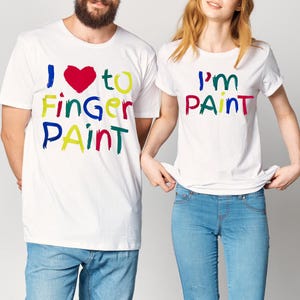May include: White t-shirts with colorful text. One shirt says "I ❤️ to Finger Paint" with a red heart. The other shirt says "I'm PAINT" in various colors. Both shirts are paired with blue jeans.