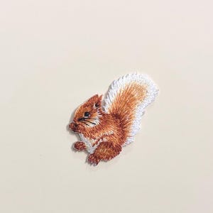 Puede incluir: Parche bordado de una ardilla en tonos marrones y blancos. La ardilla se representa de perfil, con su cola tupida y pequeñas patas. El parche es probablemente para ropa o accesorios.