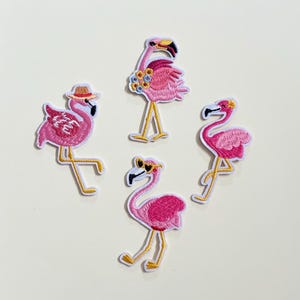 Peut inclure: Quatre écussons brodés de flamants roses dans différentes poses. Les flamants roses sont principalement roses avec des pattes et des becs jaunes. L'un porte un chapeau de paille, un autre une couronne de fleurs et un troisième des lunettes de soleil. Les écussons sont soulignés en blanc.