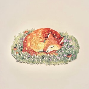 May include: Embroidered patch featuring a sleeping fox with orange and white fur, curled up on a bed of green grass and small purple flowers. Several small brown mushrooms are scattered around the fox. The patch is a whimsical, nature-themed design.