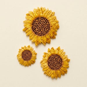 以下が含まれることがあります： さまざまなサイズの刺繍されたヒマワリのパッチが3つ。 花びらは鮮やかな黄色で、中心部は濃い茶色です。 パッチはクリーム色の背景に配置され、詳細なステッチが施されています。