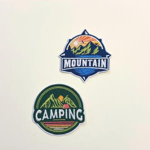 May include: Two embroidered patches with outdoor themes. One patch is a blue and white mountain scene with the word "MOUNTAIN". The other is a green and yellow camping scene with the word "CAMPING".