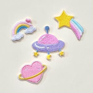 Op de afbeelding: Geborduurde patches met een regenboog, een vallende ster, een roze UFO en een roze hart met een gele ring. De patches hebben een witte rand en zijn in pastelkleuren.