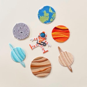 Puede incluir: Un conjunto de siete parches bordados con temas espaciales, que incluye una figura de astronauta blanca, una Tierra azul y verde, una luna gris con cráteres, un planeta rojo y naranja, un planeta azul claro con anillos, uno marrón y otro beige.
