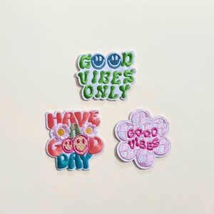 Puede incluir: Tres parches bordados con mensajes positivos. Uno dice "GOOD VIBES ONLY" en verde y azul. Otro dice "HAVE GOOD DAY" con cerezas y flores. El tercero es una flor rosa con "good vibes" en rosa.