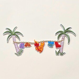 May include: An embroidered patch featuring two green palm trees connected by a gold clothesline. Hanging from the line are miniature garments including a purple patterned top, orange shorts, a red hat, an orange dress, a blue striped shirt, and red trousers.