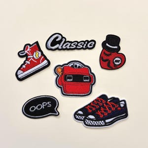 Könnte beinhalten: Eine Sammlung von aufgestickten Aufnähern. Designs umfassen einen roten High-Top-Sneaker, einen roten View-Master, ein Herz mit Zylinder und marineblaue Sneaker mit roten Schnürsenkeln. Die Aufnäher mit den Wörtern "Classic" und "Oops" sind ebenfalls enthalten.