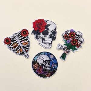 Pode incluir: Quatro patches bordados com tema de Halloween. Um é uma caixa torácica com abóboras laranja e flores vermelhas. Outro é uma caveira com uma rosa vermelha. Um terceiro é um buquê de flores com caveiras. O último é uma caveira em um vitral.