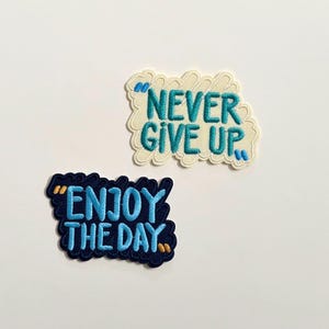 Peut inclure: Deux écussons brodés avec des citations inspirantes. L'un dit "NEVER GIVE UP" en bleu sarcelle sur fond blanc cassé. L'autre dit "ENJOY THE DAY" en bleu sarcelle sur fond bleu marine.