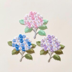May include: Three embroidered hydrangea flower patches featuring detailed stitching. One patch displays pink and white petals, another shows blue and white petals, and the third features purple and white petals. Each includes green leaves and stems.