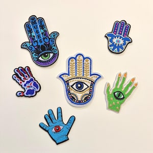 May include: A collection of six embroidered Hamsa hand patches in various colors and designs. The patches feature intricate details, including eyes, patterns, and different color schemes such as blue, green, and gold. These patches are ideal for adding a unique touch to clothing or accessories.