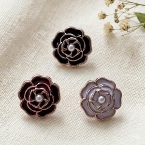 Puede incluir: Tres broches con forma de flor en negro, burdeos y gris claro. Cada broche tiene un centro de perla y un borde metálico. Los broches están dispuestos sobre una tela color crema, con flores secas en el fondo.