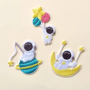 May include: Three embroidered patches featuring astronauts. One astronaut holds balloons, another fishes on a planet, and the third swings on a yellow crescent moon. Each astronaut is white with a black visor and a yellow star.