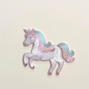 Peut inclure: Écusson brodé en forme de licorne. Le corps est blanc, la crinière et la queue sont roses, bleues et dorées, et la corne est rose. Le patch est bordé de marron clair sur fond crème.