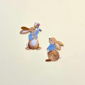 Peut inclure: Deux écussons brodés en forme de lapin. L'un des lapins tient un bouquet de fleurs, portant une veste bleue et un collier de perles. L'autre lapin porte une veste bleue et un collier de perles. Les deux lapins ont une fourrure brune et un ventre blanc.